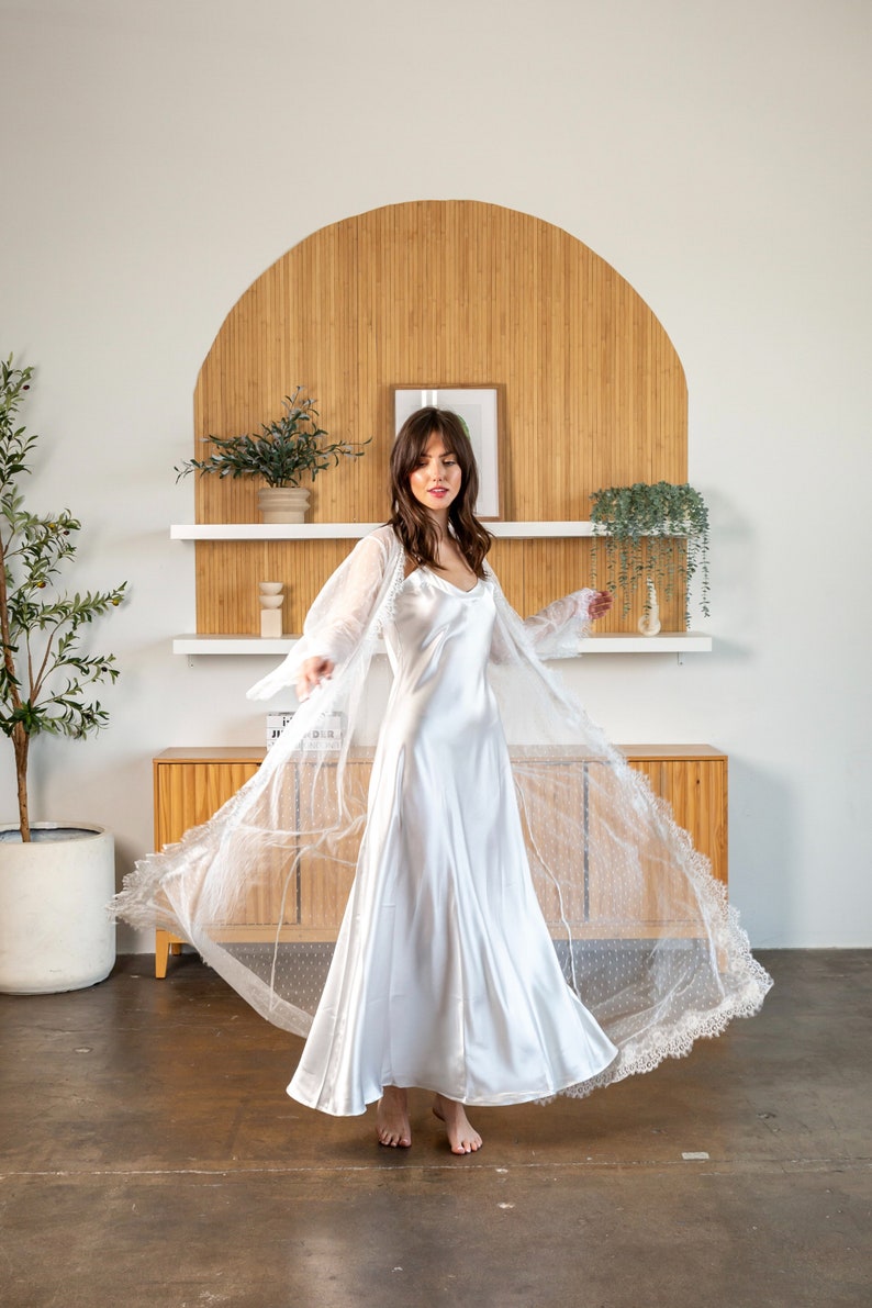 May include: A white satin slip dress with a sheer lace overlay. The dress has a V-neckline and a flowing skirt. The model is wearing the dress in a room with a wooden wall behind her.