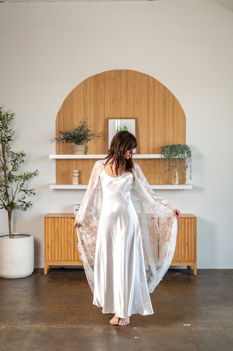 May include: A white satin slip dress with a white lace overlay. The dress is flowing and the lace is delicate. The model is standing in front of a wooden wall with shelves.