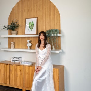 May include: A woman wearing a white lace robe over a white satin slip dress. She is sitting on a wooden cabinet with a shelf above it. The shelf has a potted plant, a sculpture, and a framed picture of a green leaf.
