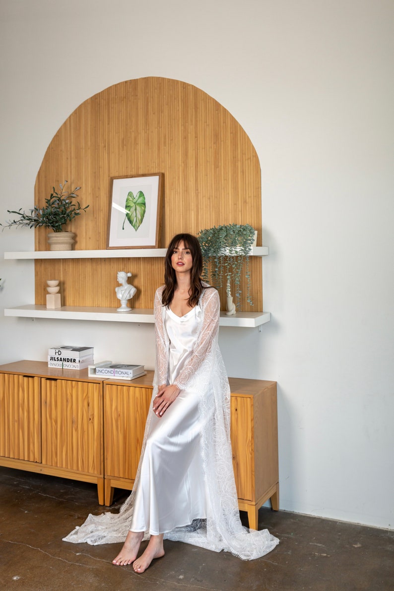 May include: A woman wearing a white lace robe over a white satin slip dress. She is sitting on a wooden cabinet with a shelf above it. The shelf has a potted plant, a sculpture, and a framed picture of a green leaf.