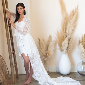 Pode incluir: Um roup&atilde;o de renda branco com mangas compridas e uma cauda longa. O roup&atilde;o tem uma faixa de cetim e uma gola alta. A modelo est&aacute; em p&eacute; em um quarto com uma escada de madeira e grama de pampa seca.