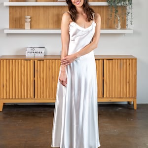 May include: A woman wearing a white satin slip dress in front of a wooden cabinet with shelves. The cabinet has a book titled "i-D JIL SANDER" on the top shelf.