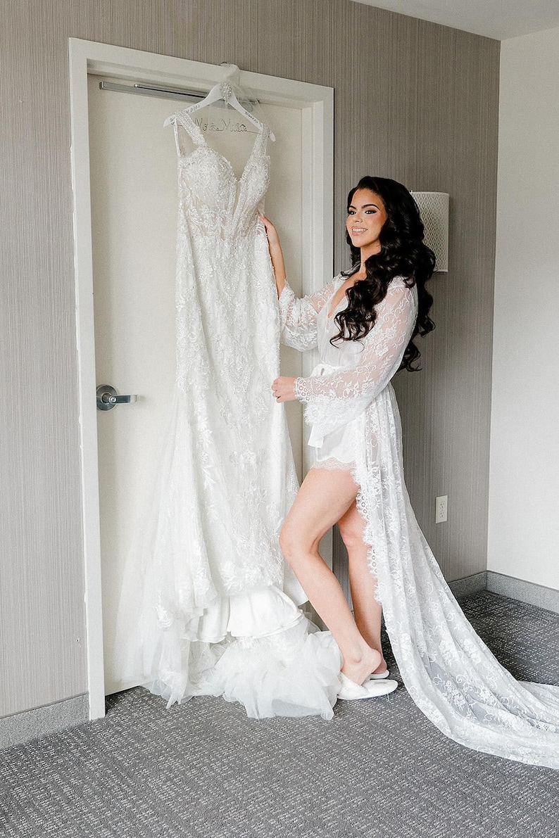 May include: A white lace wedding dress hanging on a white hanger. The dress has a sweetheart neckline and a fitted bodice. The skirt is full and flowing. The dress is displayed in a room with a white door and gray carpet.