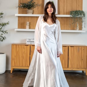 May include: A white satin slip dress with a lace trim, worn with a matching white lace robe. The model is standing in front of a wooden cabinet with a book titled "Jil Sander" on top.