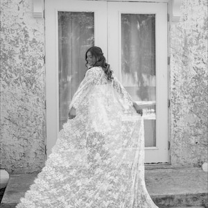 May include: A woman in a white lace dress and a long, flowing lace cape stands in front of a white door. She is smiling and looking to the side.