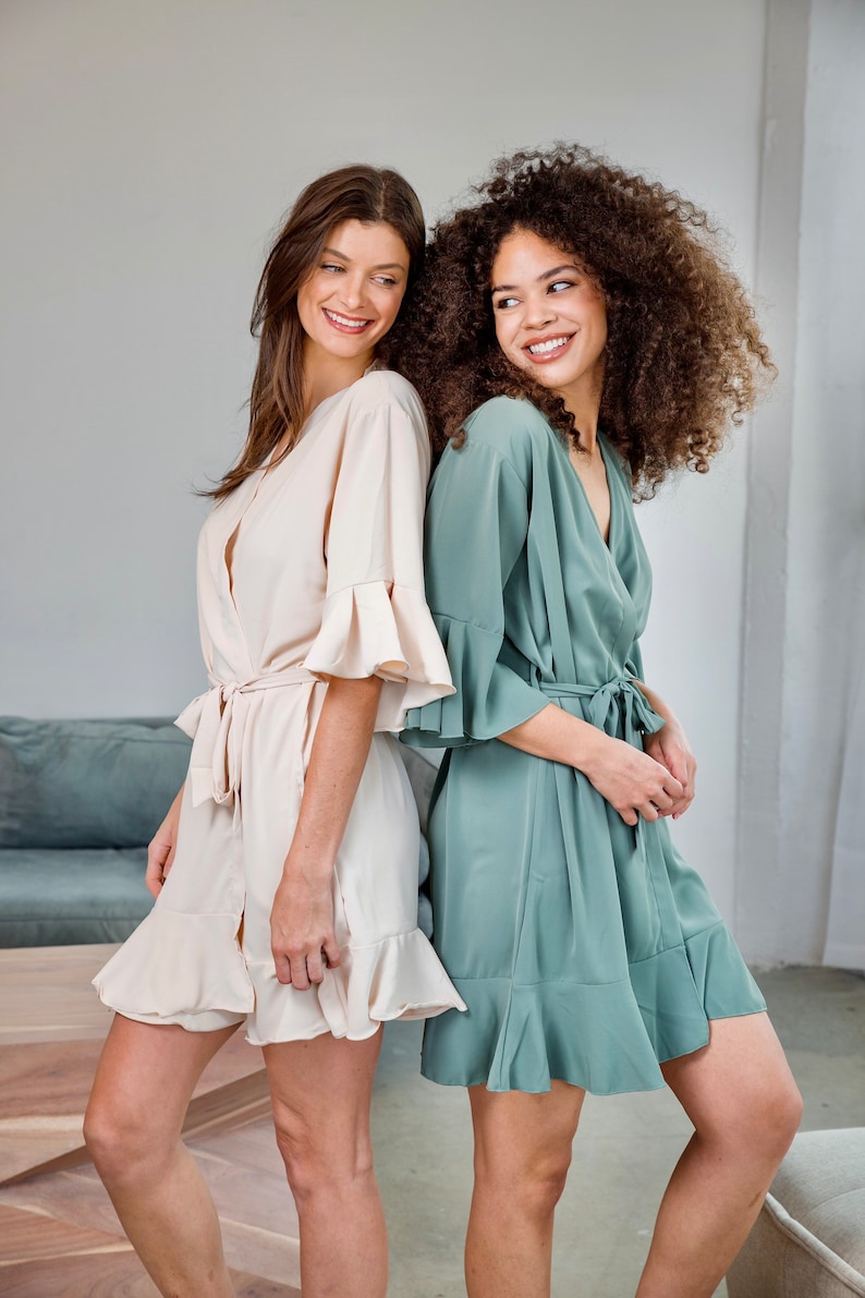 May include: Two women wearing matching robes with ruffled sleeves and hems. One robe is a light beige color, and the other is a sage green color.