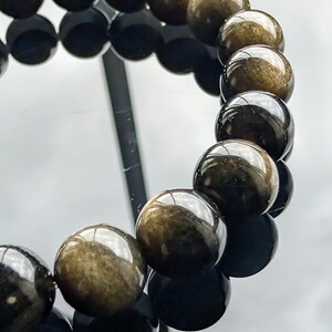May include: A bracelet featuring spherical beads with a polished, dark surface and golden flecks. The beads are arranged in a circular pattern, reflecting light and creating a shimmering effect. The bracelet is displayed on a reflective surface.