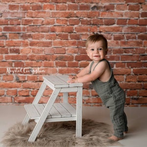 May include: A young child in a green jumpsuit stands next to a white wooden step stool. The child is smiling and looking at the camera. The background is a brick wall.