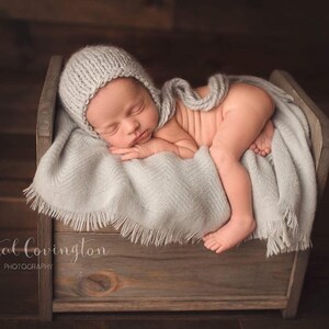 K&ouml;nnte beinhalten: Ein neugeborenes Baby in einer grauen Strickm&uuml;tze schl&auml;ft in einem Holzbettchen mit einer grauen Decke. Das Baby liegt zusammengerollt mit den Beinen unter dem K&ouml;rper.