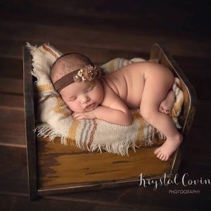 K&ouml;nnte beinhalten: Ein neugeborenes Baby schl&auml;ft auf einem kleinen Holzbett mit einer braunen und wei&szlig;en Decke. Das Baby tr&auml;gt ein braunes Stirnband mit einem Blumenakzent.