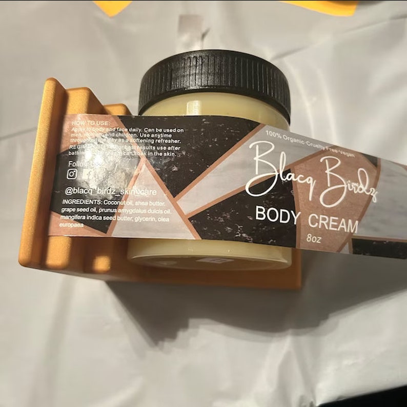 May include: A glass jar of body cream with a black lid and a label that says "Blacq Birds Body Cream 8oz". The label is black and gold with geometric shapes and the text "100% Organic Cruelty Free Vegan".