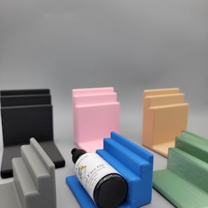 May include: Five different colored tiered display stands made of plastic. The stands are in black, pink, beige, blue, and green. A small bottle of face oil is sitting on the blue stand.