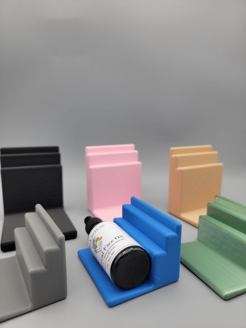 May include: Five different colored tiered display stands made of plastic. The stands are in black, pink, beige, blue, and green. A small bottle of face oil is sitting on the blue stand.