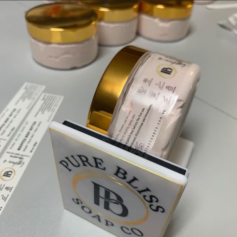 May include: A white square tile with gold trim and black text that reads "PURE BLISS SOAP CO" with a stylized black letter "B" in the center. A jar of body butter with a gold lid and a white label with black text is sitting on the tile.