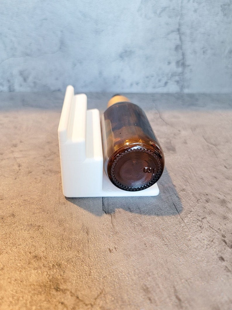 May include: White ceramic bottle holder with a brown glass bottle resting inside. The bottle holder has three steps and is designed to hold a bottle upright.