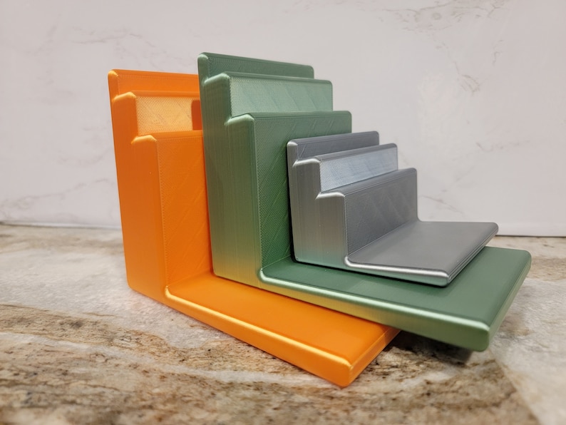 May include: A set of five stacked, angled shelves made of 3D printed plastic. The shelves are in shades of orange, green, and silver.