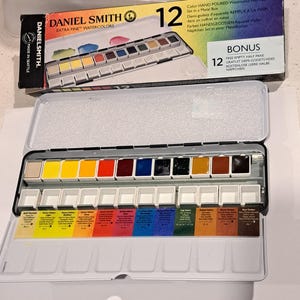 May include: A Daniel Smith Extra Fine Watercolours set in a metal box. The set includes 12 colours in half pans, with a bonus of 12 empty half pans. The box is labelled "Made in Seattle."