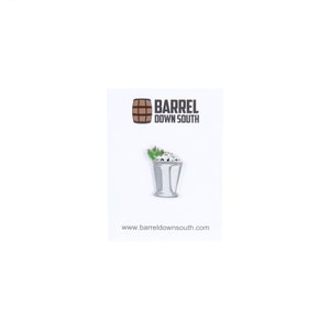 May include: A white enamel pin with a silver outline of a mint julep cocktail. The pin is on a white card with the text "Barrel Down South" and a brown barrel logo. The website address "www.barreldownsouth.com" is printed below the logo.