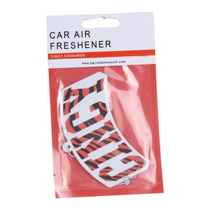 Peut inclure: Un désodorisant pour voiture dans un emballage plastique transparent. Le désodorisant est blanc avec le mot "CINCY" en rayures tigrées orange et noir. L'emballage indique "CAR AIR FRESHENER" et "CINCY CINNAMON".