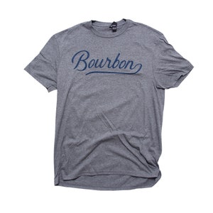 May include: A heather gray t-shirt with the word "Bourbon" in a navy blue script across the chest. The shirt has short sleeves and a crew neck. The fabric appears soft and slightly wrinkled.