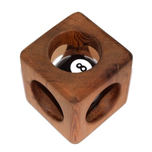 Può includere: Un porta-palla da biliardo a forma di cubo in legno. Il supporto è in legno marrone e presenta tre aperture circolari. All'interno del supporto c'è una palla nera e bianca con il numero 8. La venatura del legno è visibile.