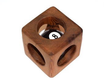 May include: A wooden cube-shaped billiard ball holder. The holder is made of brown wood and has three circular openings. Inside the holder is a black and white eight ball. The wood grain is visible on the surface.