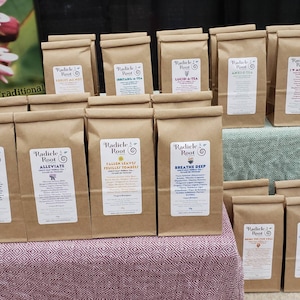 May include: A display of brown paper bags with labels for different herbal teas. The labels feature the brand name "Radicle Root" and the name of each tea blend. The tea blends include "Stimulate", "Alleviate", "Fallen Leaves", "Breathe Deep", "Forget Me Not", "Irritaris-A-Tea", "Lucid-A-Tea", "Amre-A-Tea", "I W/ My Uterus", "Brown Rice Paw Chai", and "Coat A Throat".