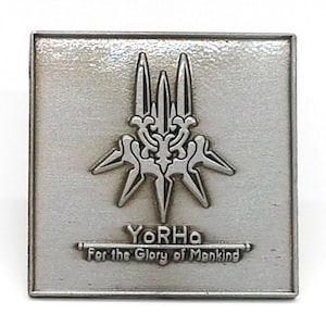 Könnte beinhalten: Quadratischer, silberfarbener Anstecker mit einem stilisierten Emblem mit drei vertikalen Elementen und sternartigen Spitzen. Der Text "YoRHa For the Glory of Mankind" ist darunter eingraviert. Der Anstecker hat eine strukturierte Oberfläche.