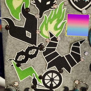以下が含まれることがあります： ファンタジーをテーマにした刺繍パッチとキーホルダーのコレクション。パッチは、ドラゴン、炎、その他のファンタジー要素の黒と緑のデザインが特徴です。キーホルダーは、ネオンの輝きを持つ稲妻のデザインです。