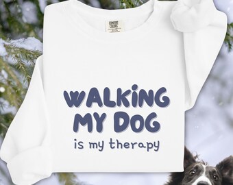 Puede incluir: Sudadera blanca de cuello redondo con el texto azul "WALKING MY DOG is my therapy". La sudadera está hecha de un material suave y es perfecta para los amantes de los perros. La cara de un perro es visible en la esquina inferior derecha.