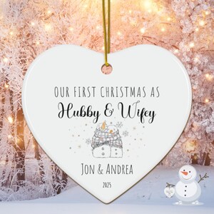 Peut inclure: Ornement de Noël en forme de cœur blanc avec le texte "OUR FIRST CHRISTMAS AS Hubby & Wifey". Il présente un couple de bonhommes de neige dans une écharpe, avec "JON & ANDREA" et l'année "2025" en dessous. Une ficelle dorée est attachée pour l'accrocher. Environ 10 cm de large.