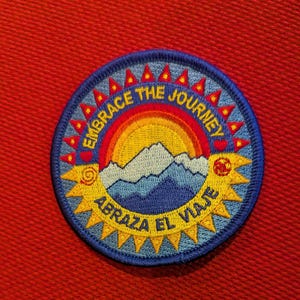 May include: Embroidered patch with a blue border and the text "EMBRACE THE JOURNEY" and "ABRAZA EL VIAJE". The patch features a colorful design of a rainbow, mountains, and sun. The patch is round and has a yellow and red border.