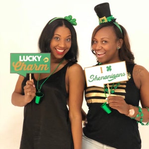 Puede incluir: Dos mujeres con atuendos temáticos del Día de San Patricio en verde y dorado. Una mujer sostiene un cartel que dice "Lucky Charm" y la otra sostiene un cartel que dice "I ♥ Shenanigans".