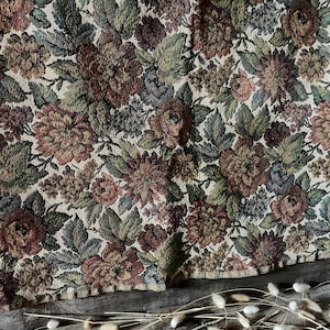 Puede incluir: Una tela de tapiz tejida con un denso patrón floral en tonos óxido, verde salvia, oliva y azul apagado. El textil muestra una disposición repetitiva de flores en flor y follaje detallado sobre un fondo de color beige claro.