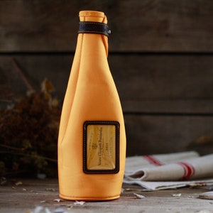 Puede incluir: Una funda para botella de tela texturizada de color amarillo brillante con una etiqueta ribeteada en cuero marrón oscuro con el texto Veuve Clicquot Ponsardin Brut. La funda incluye una correa de cuero marrón oscuro a juego con un cierre de metal.