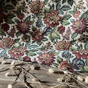Peut inclure: Un tissu tissé présentant un motif floral dense avec des fleurs rouge foncé, vert sourd et bleu sur un fond crème. Le textile présente un tissage texturé rappelant la tapisserie avec des détails botaniques complexes sur toute la surface.