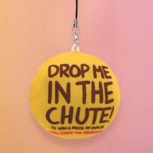 May include: A yellow circular charm with the text "DROP ME IN THE CHUTE!" in brown. The charm is attached to a black string with a silver-colored clasp. The bottom of the charm has the text "TO WIN A PRIZE ON DISPLAY CALL STAFF FOR AS".