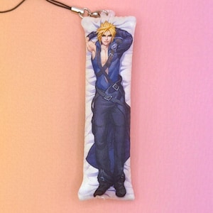 May include: A rectangular charm featuring a character with spiky blonde hair, wearing a blue and black outfit, and lying on a pillow. The charm has a black strap attached to a silver clasp. The background is a gradient of pink and peach.