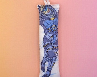 May include: A rectangular, white plush charm featuring a blue and yellow illustration of a futuristic space suit. The charm is attached to a black lanyard with a silver-colored clasp. The background is a gradient of pink and peach.