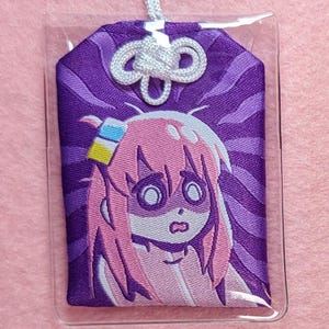 Puede incluir: Un amuleto de tela morado y blanco con un personaje de dibujos animados con cabello rosado y una expresi&oacute;n de sorpresa. El amuleto est&aacute; en una caja de pl&aacute;stico transparente con una cuerda blanca atada.
