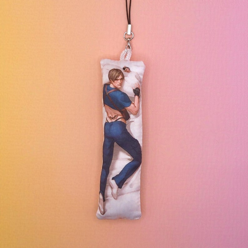 May include: A rectangular charm with a character illustration in a relaxed pose, wearing a blue top and trousers. The charm is attached to a black cord and silver-toned hardware, with a white pillow background.
