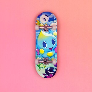 May include: A colorful finger skateboard featuring cartoon characters in shades of blue, yellow, and green against a sky-blue background. The skateboard has white wheels and silver metal trucks, set against a pink backdrop.