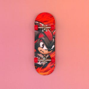 May include: A red and black finger skateboard featuring a cartoon character design. The skateboard has red wheels and silver axles. The character is depicted with black and red coloring against a fiery background.