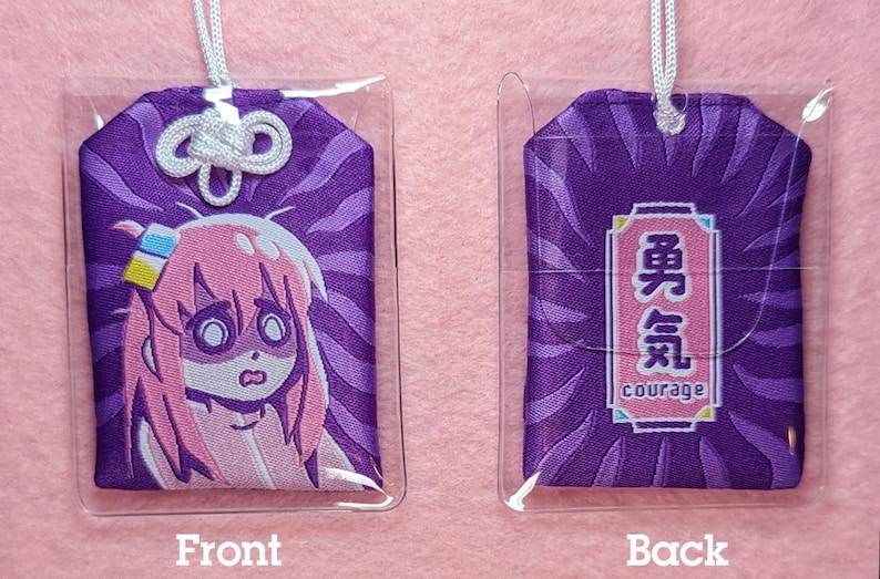 Puede incluir: Encanto de tela morado con una cuerda blanca atada. La parte delantera presenta una ilustraci&oacute;n de dibujos animados de una ni&ntilde;a de pelo rosa con una expresi&oacute;n de sorpresa. La parte trasera presenta un rect&aacute;ngulo morado y rosa con el texto "勇気" en japon&eacute;s y "Courage" en ingl&eacute;s.