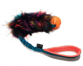 May include: A colorful, multi-colored, fuzzy dog toy with a bright orange rubber ball on the end. The toy has a teal blue rope handle with a black label that says "Sivas" and a star.