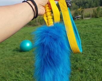 Puede incluir: Un juguete para perros azul y amarillo con una cola larga y esponjosa azul y un mango de cuerda amarillo. El juguete está sostenido en una mano con una pulsera negra.
