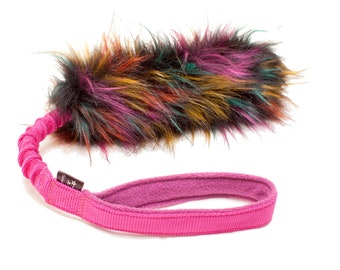 May include: A pink and black dog toy with a long, colorful fur tail. The toy has a pink handle with a black label.