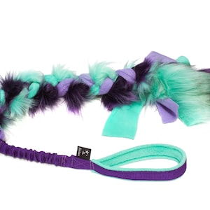 Op de afbeelding: Een gevlochten hondenspeeltje in paars, teal en zwart. Het speeltje heeft een zachte, harige textuur en een handvat met een bijpassend kleurenschema. Het handvat is met een paarse elastische band aan het gevlochten gedeelte bevestigd. Voor interactief spelen.