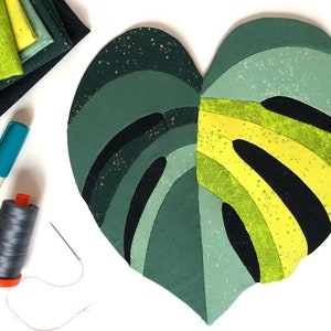Puede incluir: Una hoja de monstera de tela verde y amarilla, hecha con cuadrados de tela de patchwork, con detalles de purpurina dorada. La hoja está sobre una superficie blanca con materiales de costura, incluyendo un carrete de hilo gris, una aguja y un marcador azul.