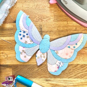 May include: A blue and purple butterfly-shaped fabric applique with a pink and white patterned design. The applique is on a wooden surface with sewing supplies.